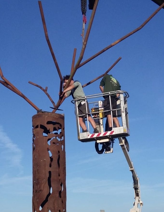 Twee mannen werken aan de installatie van een unieke stalen boom vanaf een hoogwerker, met een blauwe lucht op de achtergrond.